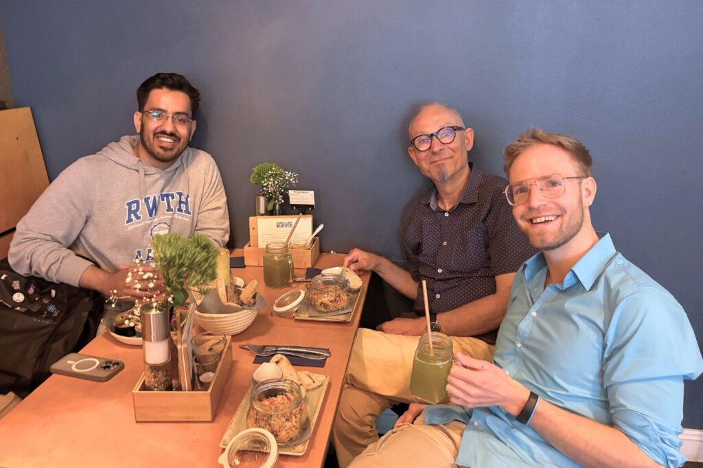 Kunal trägt ein RWTH-Hoodie und sitzt gegenüber von Helmut, einem verschmitzten älteren Herren mit Goatie und Brille, sowie Oliver im hellblauen Hemd in einem Lokal beim Essen.